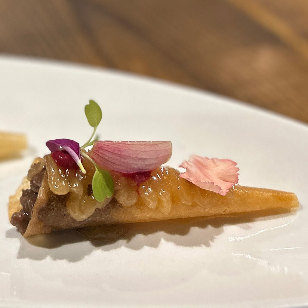 Se trata de un atuténtico festín visual y culinario. Un singular bocado, explosión de sabores y colores cuidadosamente combinados. Un canutillo relleno de tierna carne de vaca combinado con un praliné de cebolla y remolacha picante, coronado con encurtidos y pétalos de flor comestibles.